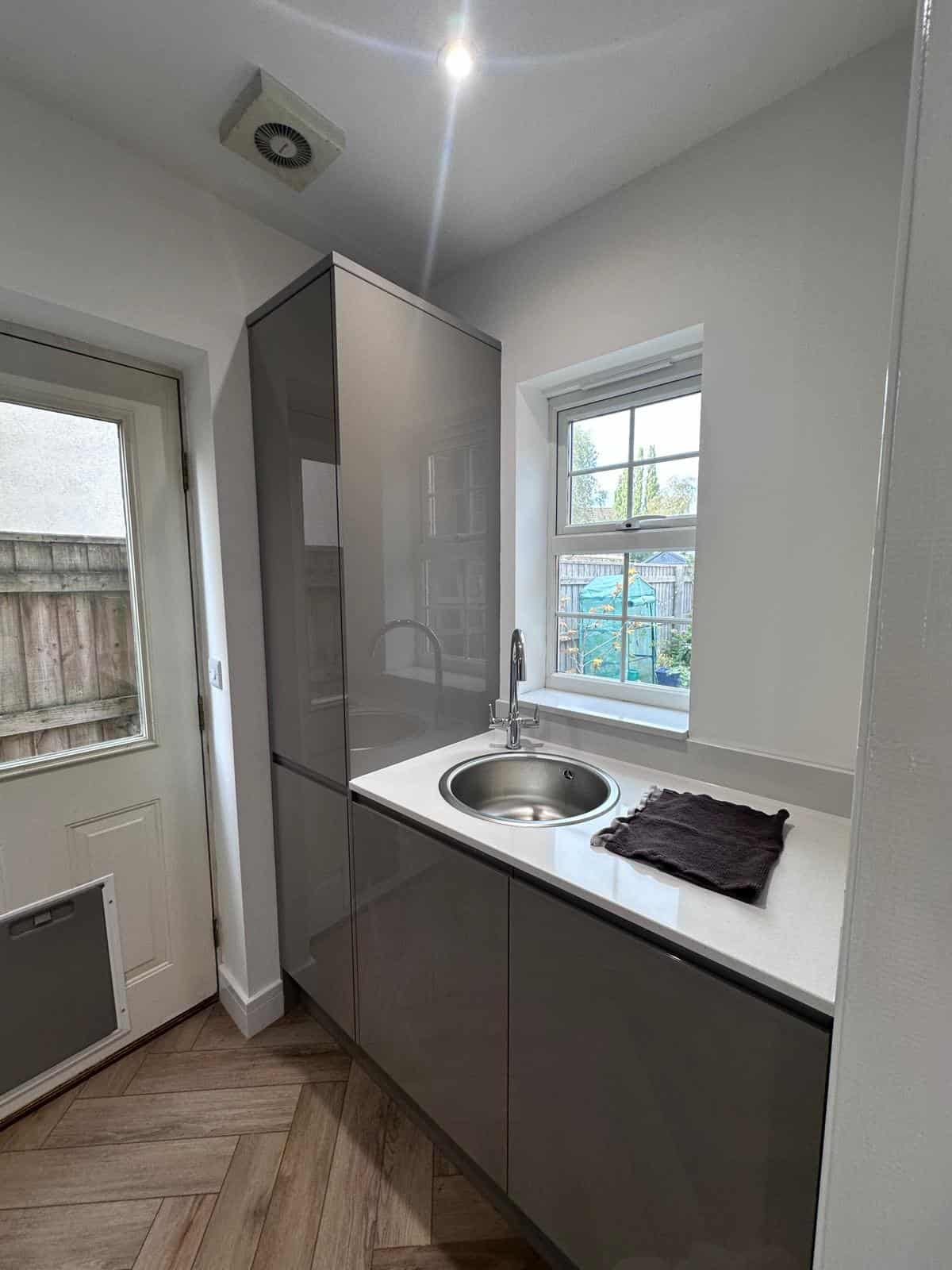 Modern grey kitchen units with stainless steel sink and white countertop, featuring a window and door.