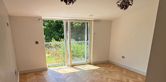 Bright empty living room with wooden flooring and large glass doors leading to balcony.
